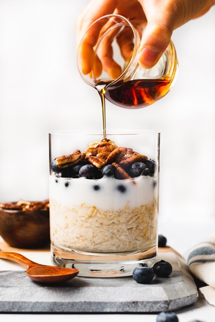 A hand pours honey from a small glass container onto a layered parfait in a clear glass. The nutritious parfait contains oats, yogurt, blueberries, and pecans. A wooden spoon, scattered blueberries, and a bowl of pecans are nearby on a light-colored surface.
