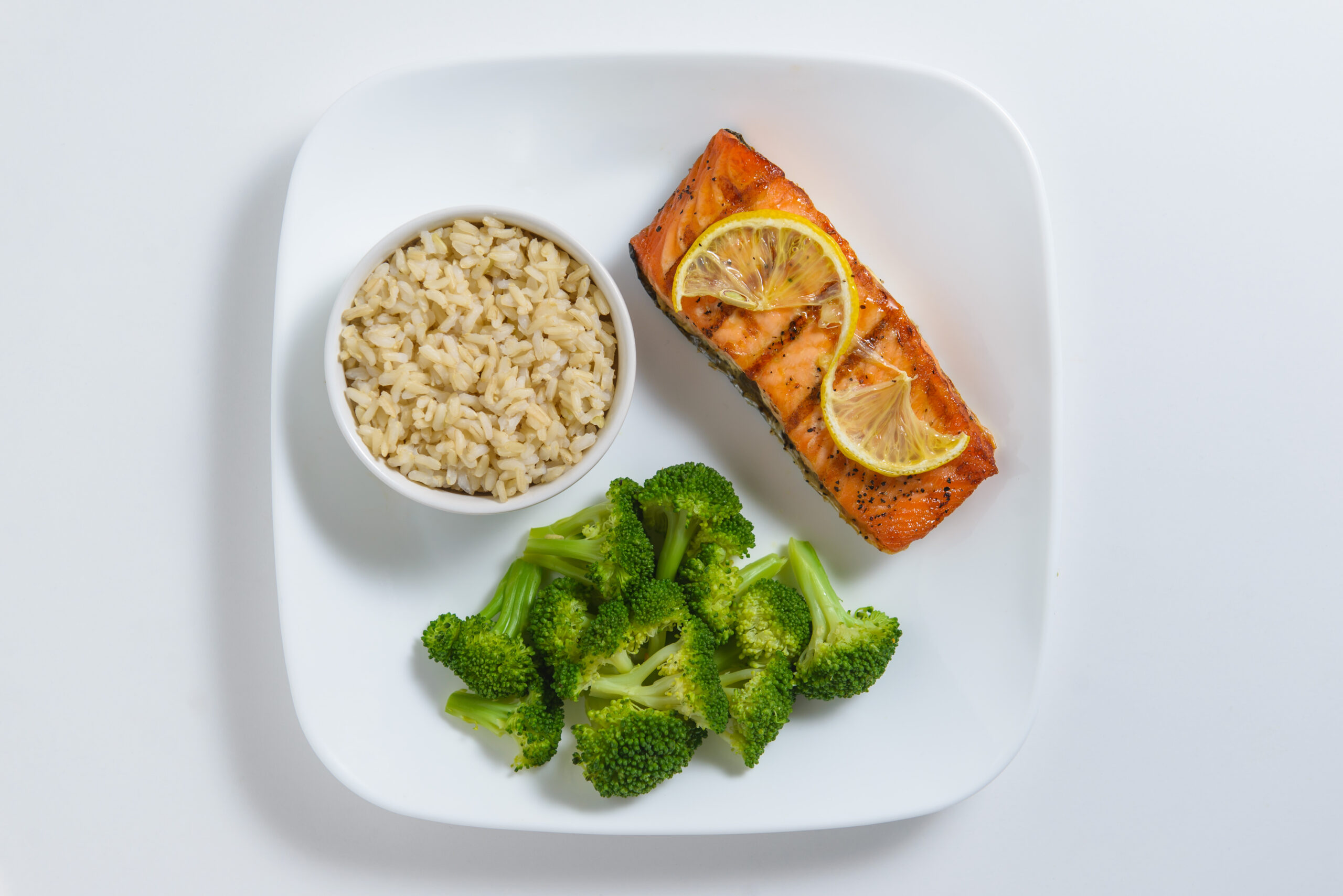 A white square plate showcases a nutritious meal with a serving of brown rice in a small bowl, a piece of baked salmon topped with two lemon slices, and steamed broccoli florets. The arrangement is neat, with the salmon placed next to the bowl of rice and the broccoli below them.