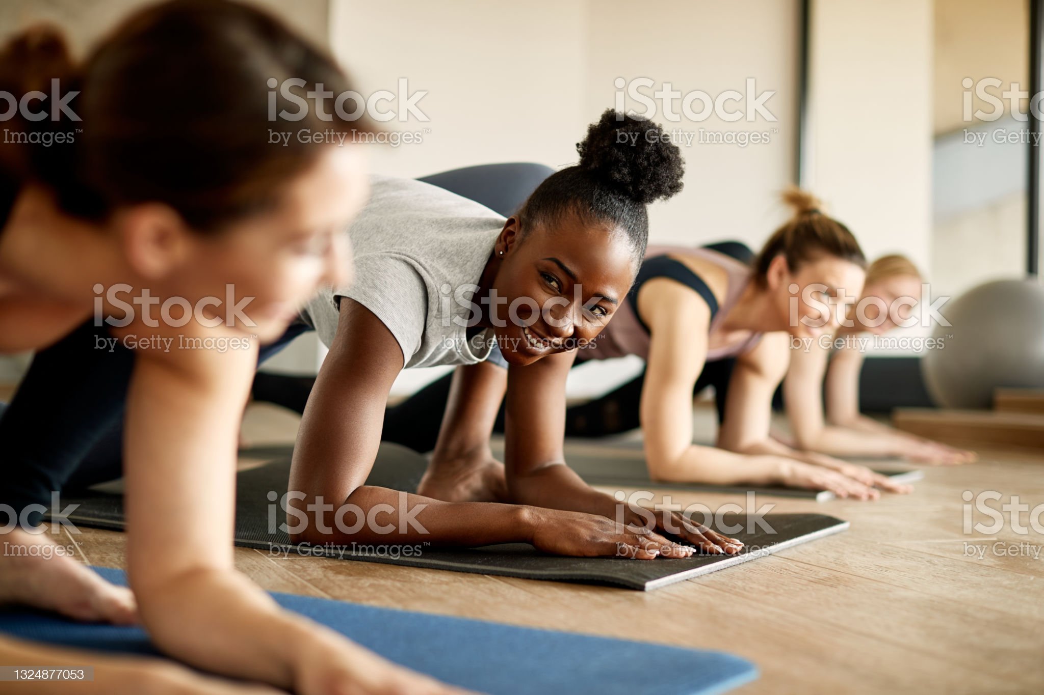 Four women are in a workout class performing plank exercises on yoga mats. They are all aligned in a row, focused on maintaining their plank positions. The woman in the forefront is smiling while looking ahead. The background is a well-lit, wooden-floored room.