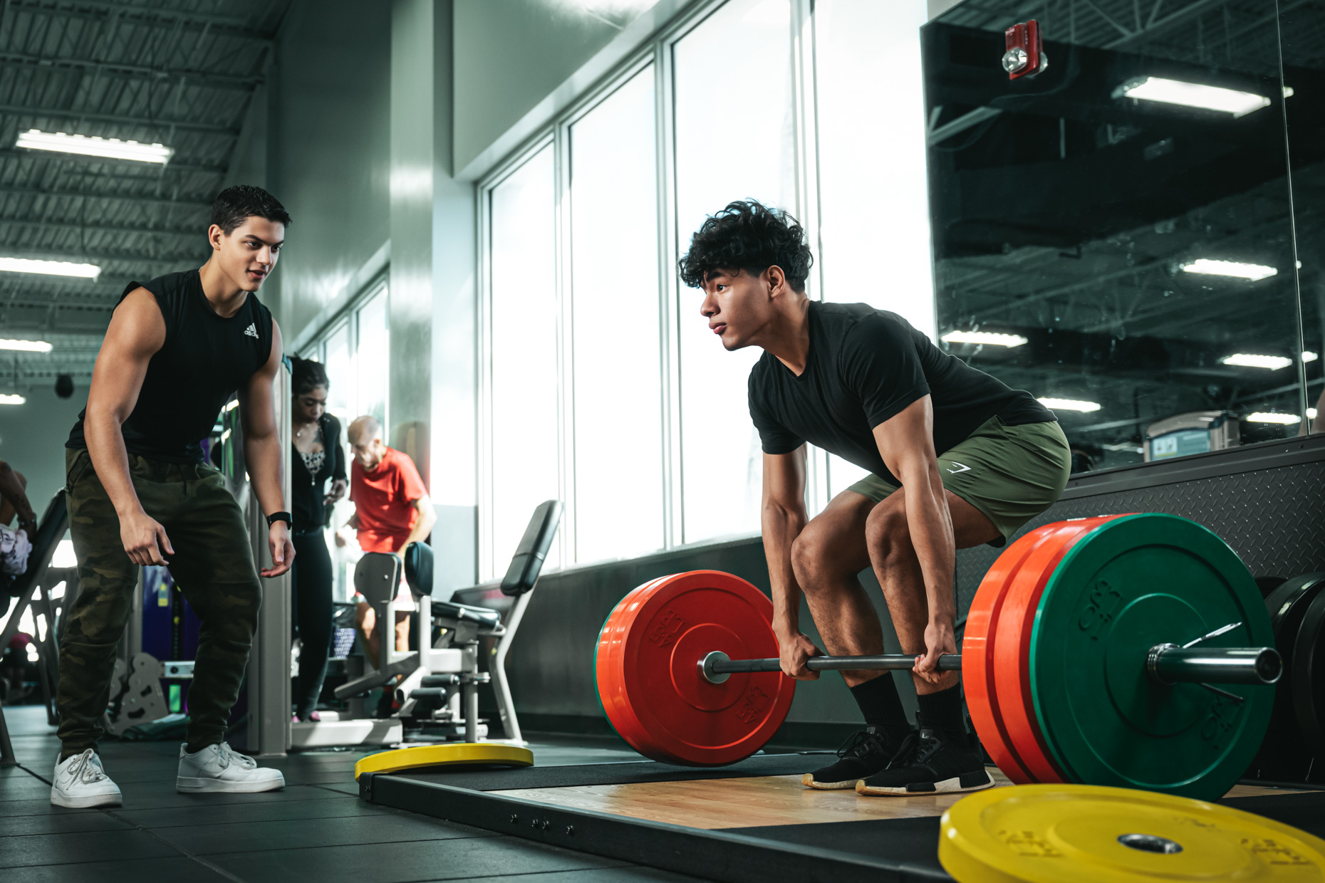 A gym with a row of black dumbbells on a rack in the foreground. In the background, a person with blonde hair wearing a black tank top bends over while lifting weights. Several other exercise machines and equipment are visible, providing everything needed for an effective beginner gym workout. The room is well-lit.