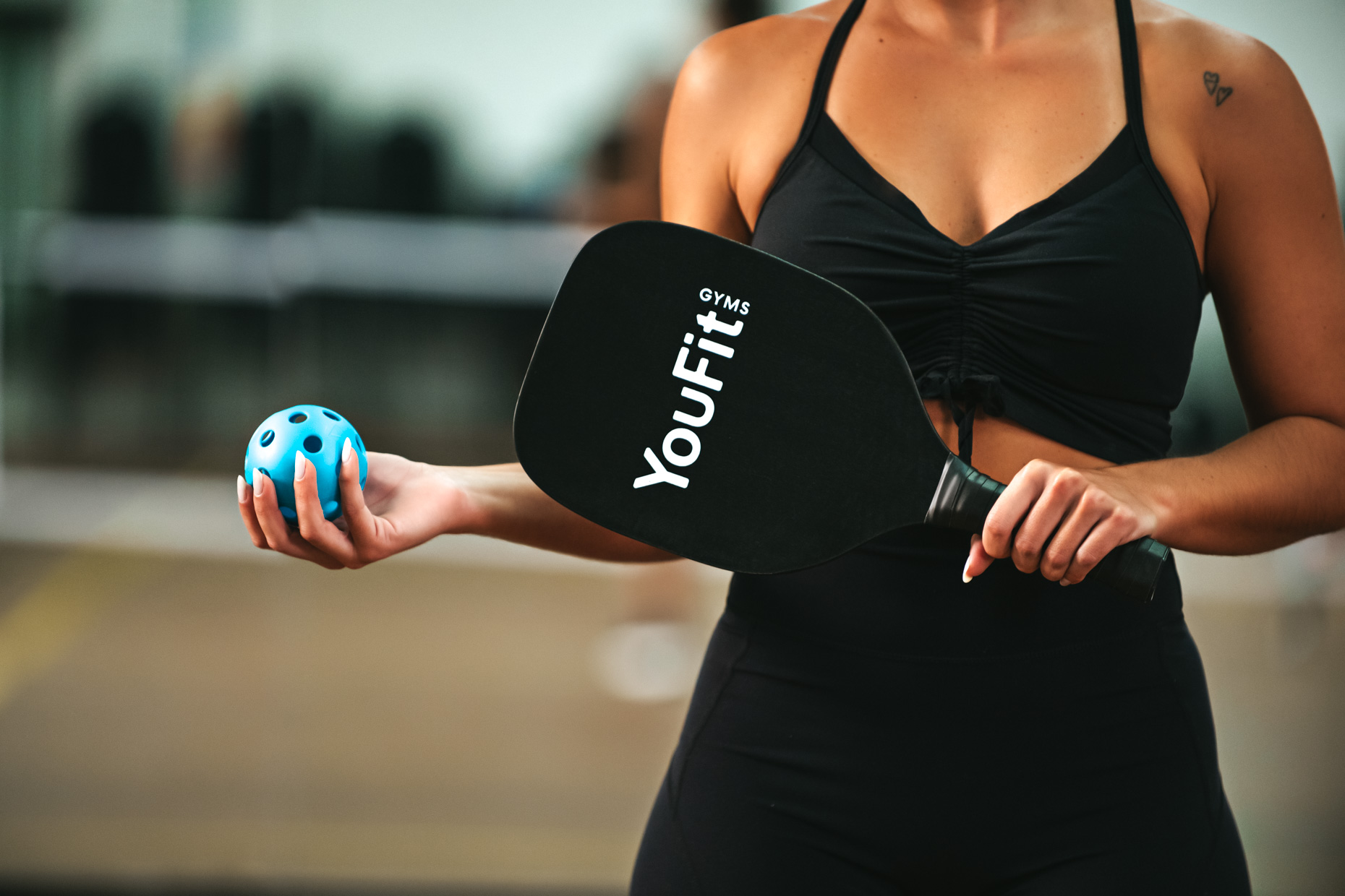 A person wearing a grey shirt, orange shorts, and knee pads is playing pickleball indoors at Youfit Gyms. They are focused on hitting the ball with a paddle, and a pickleball net is visible in the background.