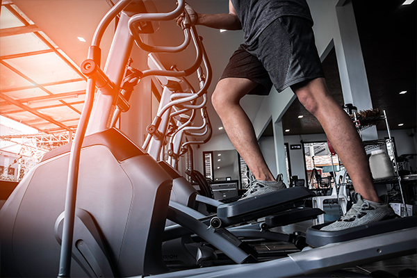 A person in athletic wear is using an elliptical machine in a gym, showcasing one of the effective cardio workouts you can do at the gym. The focus is on their legs and the exercise equipment. The well-lit gym has other exercise machines visible in the background.