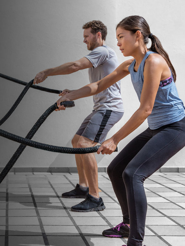 Two people are engaged in a battle ropes exercise. A woman in a blue tank top and black leggings is on the right, and a man in a gray T-shirt and shorts is on the left. They are outdoors on a checkered floor, focused on their workout, embodying the energy you’d find at Youfit Gyms.