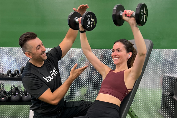 A fitness trainer assists a woman who is performing a dumbbell bench press exercise in a gym. The woman, wearing a maroon sports top, black leggings, and wireless earbuds, smiles as she works out. The background includes a dumbbell rack and metallic wall panel. It's part of her free beginner workout routine.