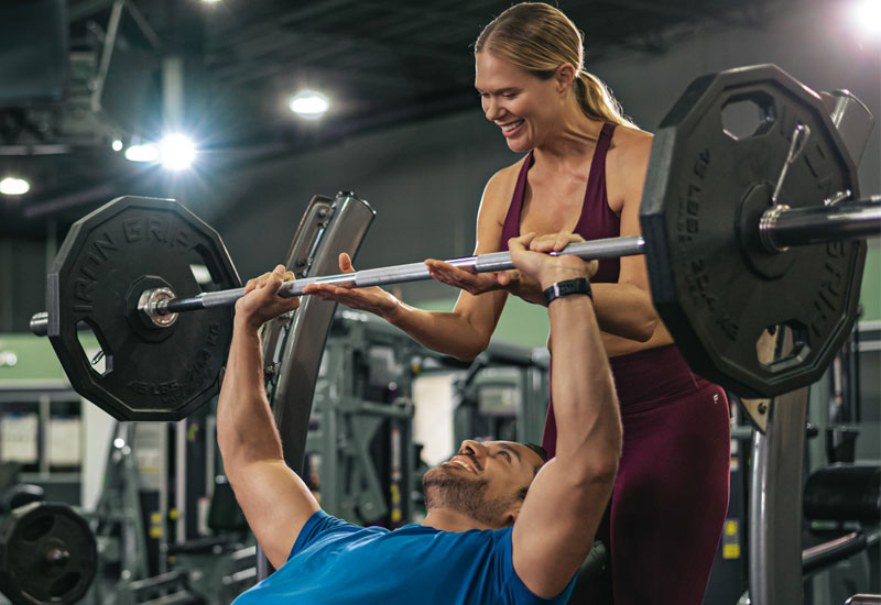 A man in a blue shirt is lying on a bench press at YouFit Gyms Kendall 112th St, lifting a weighted barbell. A woman in red athletic wear stands behind him, spotting and encouraging him. They are in a brightly lit gym with various exercise equipment in the background.