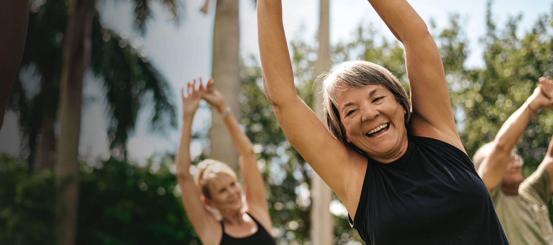 A group of people practicing yoga outdoors with palm trees in the background. The focus is on an older woman in a black tank top, smiling and stretching her arms upwards, cherishing the benefits of old memberships. Other participants are visible behind her. The sky is clear and the scene is bright and cheerful.