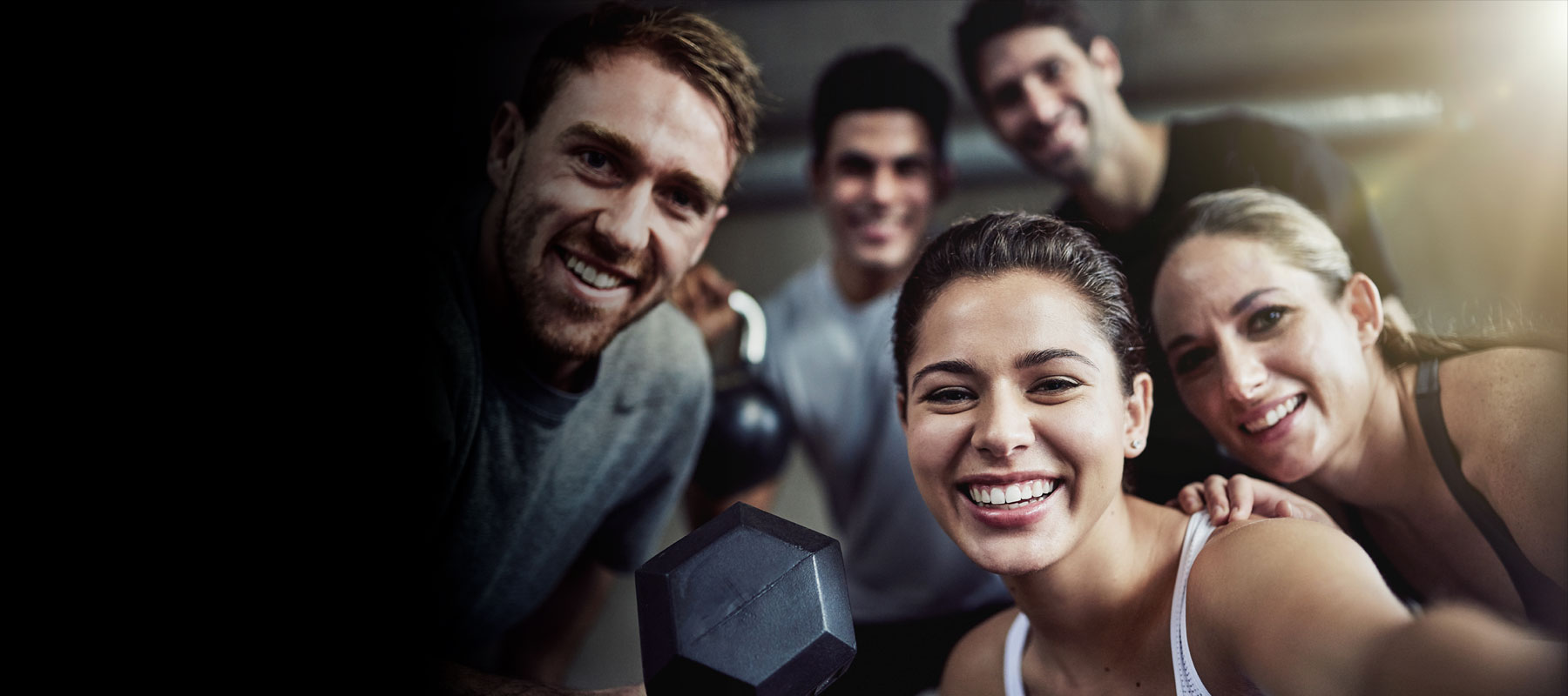 A group of five people are smiling and posing for a selfie at Youfit Gyms. They are holding a dumbbell and appear happy and energetic. The background is dimly lit, but the individuals are well-lit and in focus.