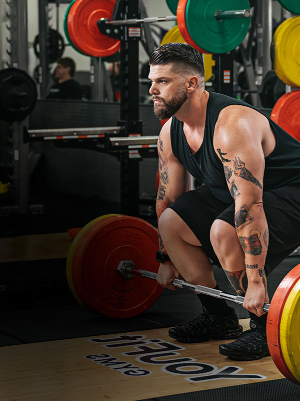 A muscular man with tattoos, wearing a sleeveless black shirt and black shorts, is preparing to lift a barbell loaded with colorful weight plates at the gym. He is in a squat position, holding the barbell with both hands, concentrating on the lift—perfecting his fitness routine after getting his gym membership.