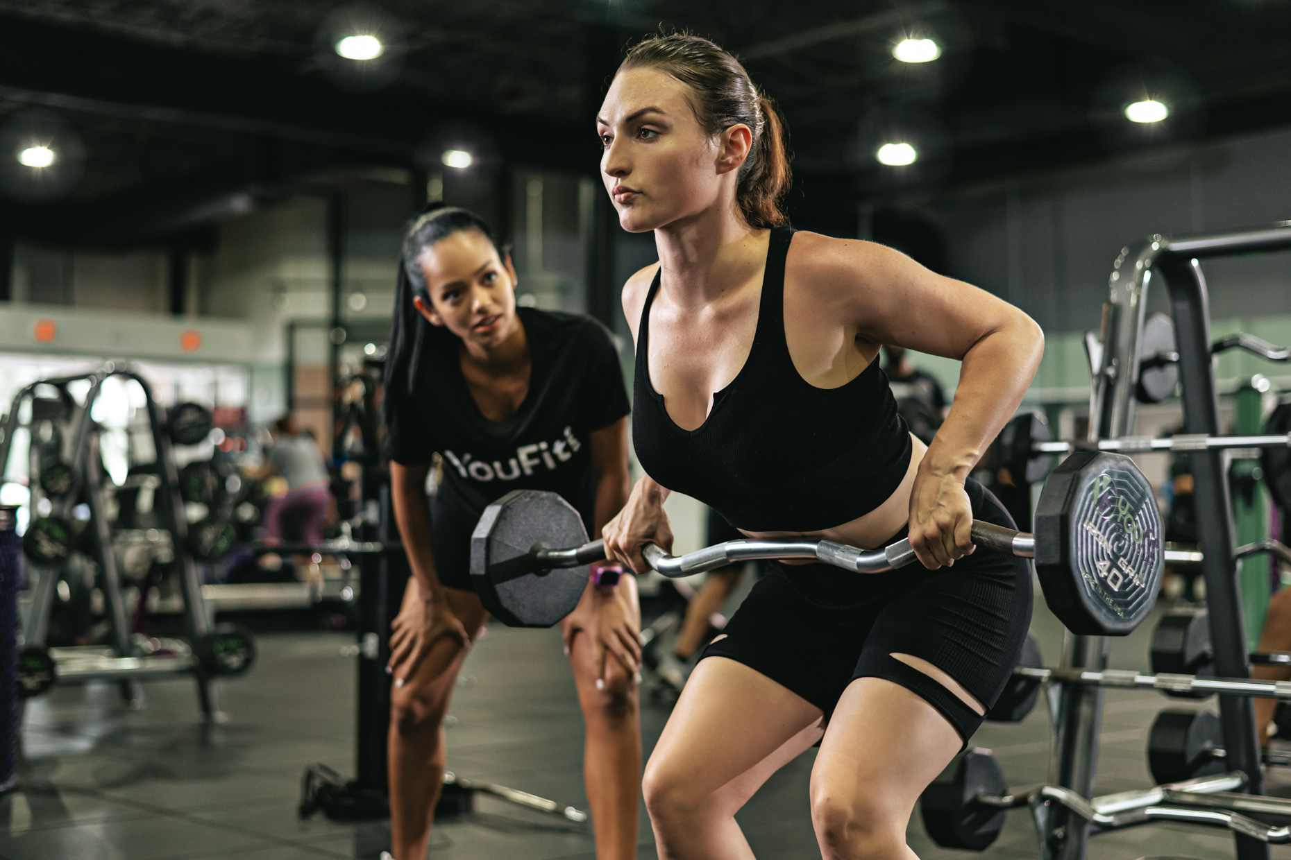 A woman in athletic wear is performing a barbell bent-over row exercise in a gym. A fitness instructor watches her closely, providing guidance. The gym, buzzing like a back-to-school season, is equipped with various exercise machines and other people can be seen working out in the background.