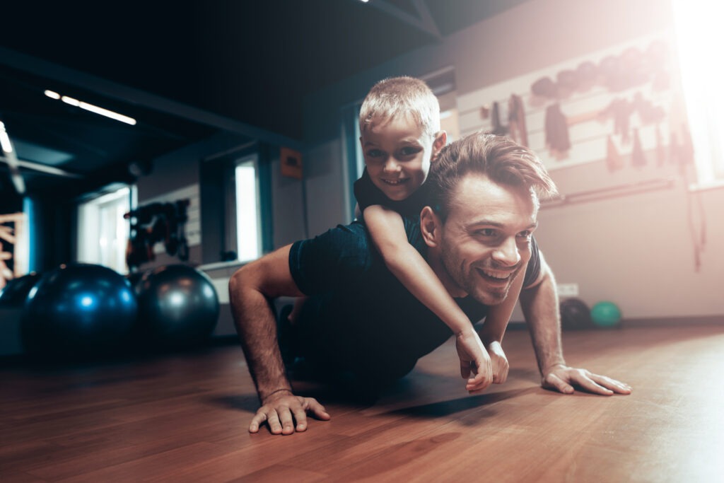 A smiling father doing pushups, weight loss training in a gym with his happy son on his back.
