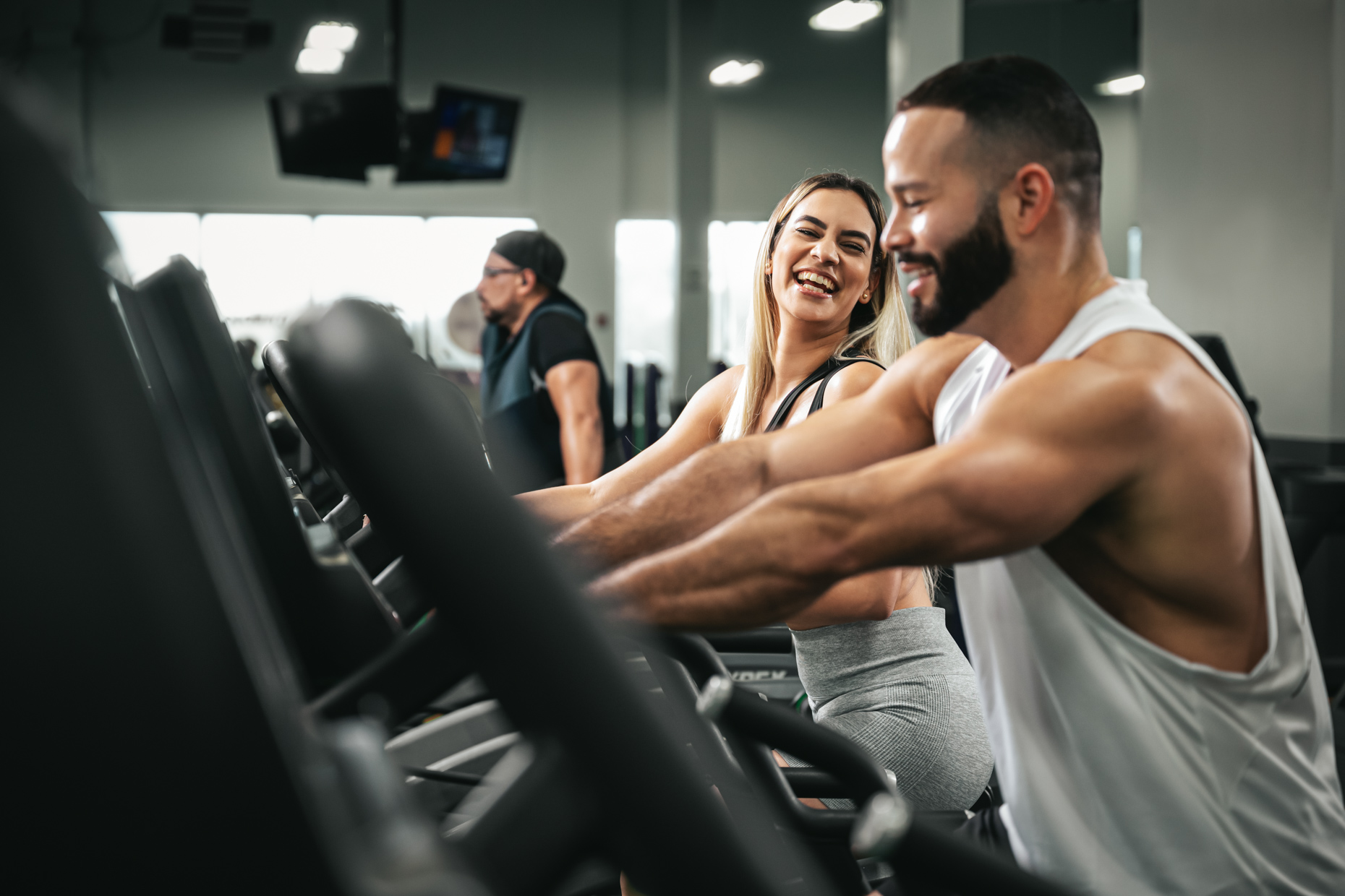 A man and woman smile while enjoying their cardio fitness routine on treadmills at a gym. The woman wears a light gray sports bra and leggings, while the man sports a white tank top. Another person jogs on a treadmill in the background, dressed in a black hat and shirt.