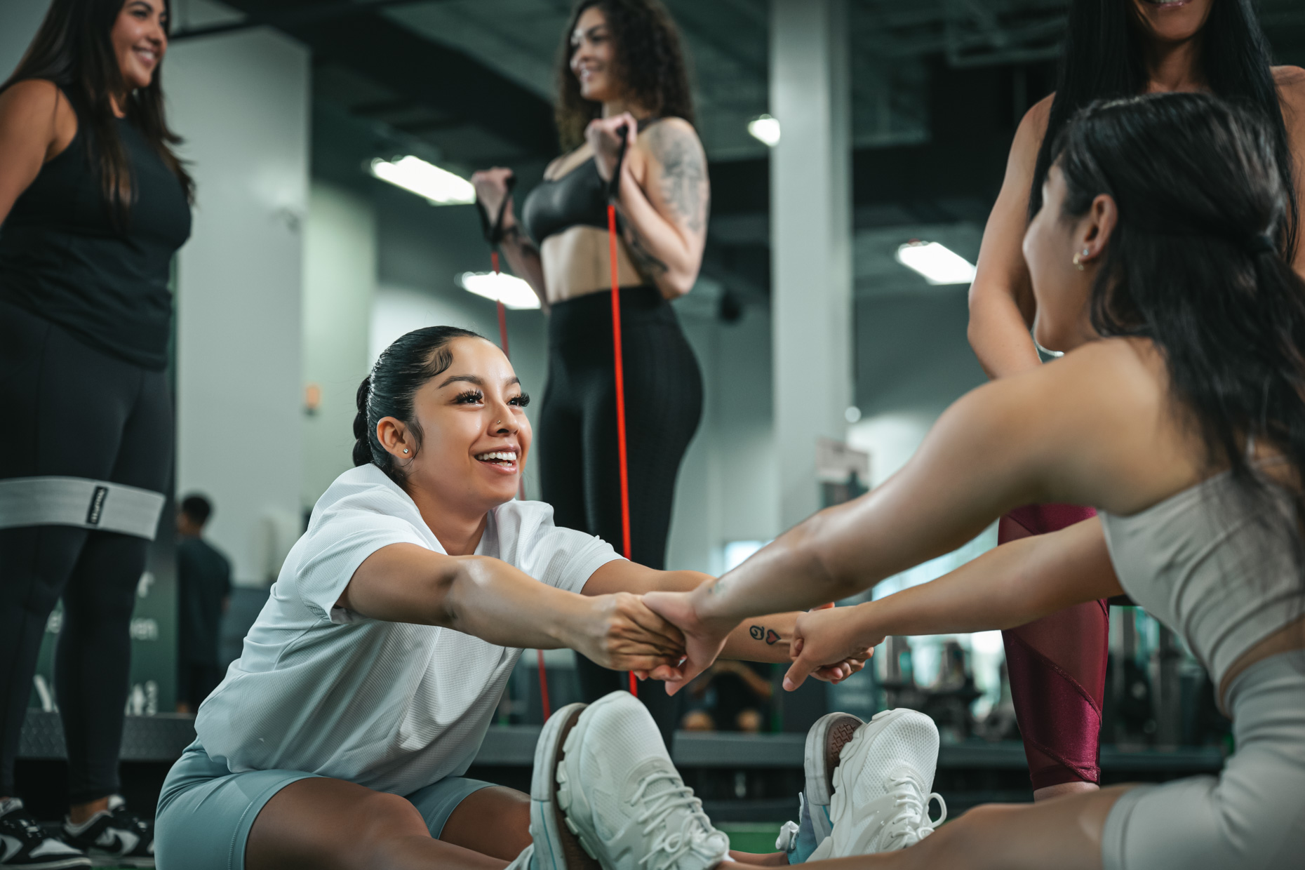 A group of women exercising in a gym cultivates healthy habits. Two women in the foreground are smiling and stretching with each other on the floor, holding hands, while others in the background exercise with resistance bands and interact with each other. The setting is a modern gym.