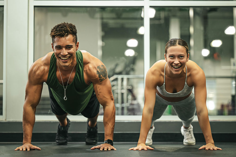 A man and a woman are performing push-ups side by side in a gym, both smiling and seeming to enjoy their workout, perhaps taking a break from babysitting duties. The gym background includes various exercise equipment and large windows. The man is wearing a green tank top and the woman a gray sports bra.