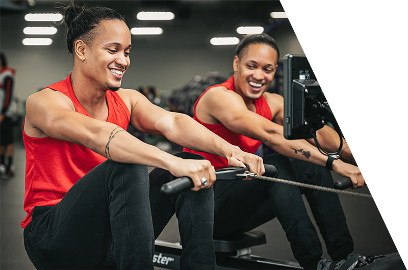 Fitness training - Two happy individuals in red sleeveless shirts using rowing machines at the gym.