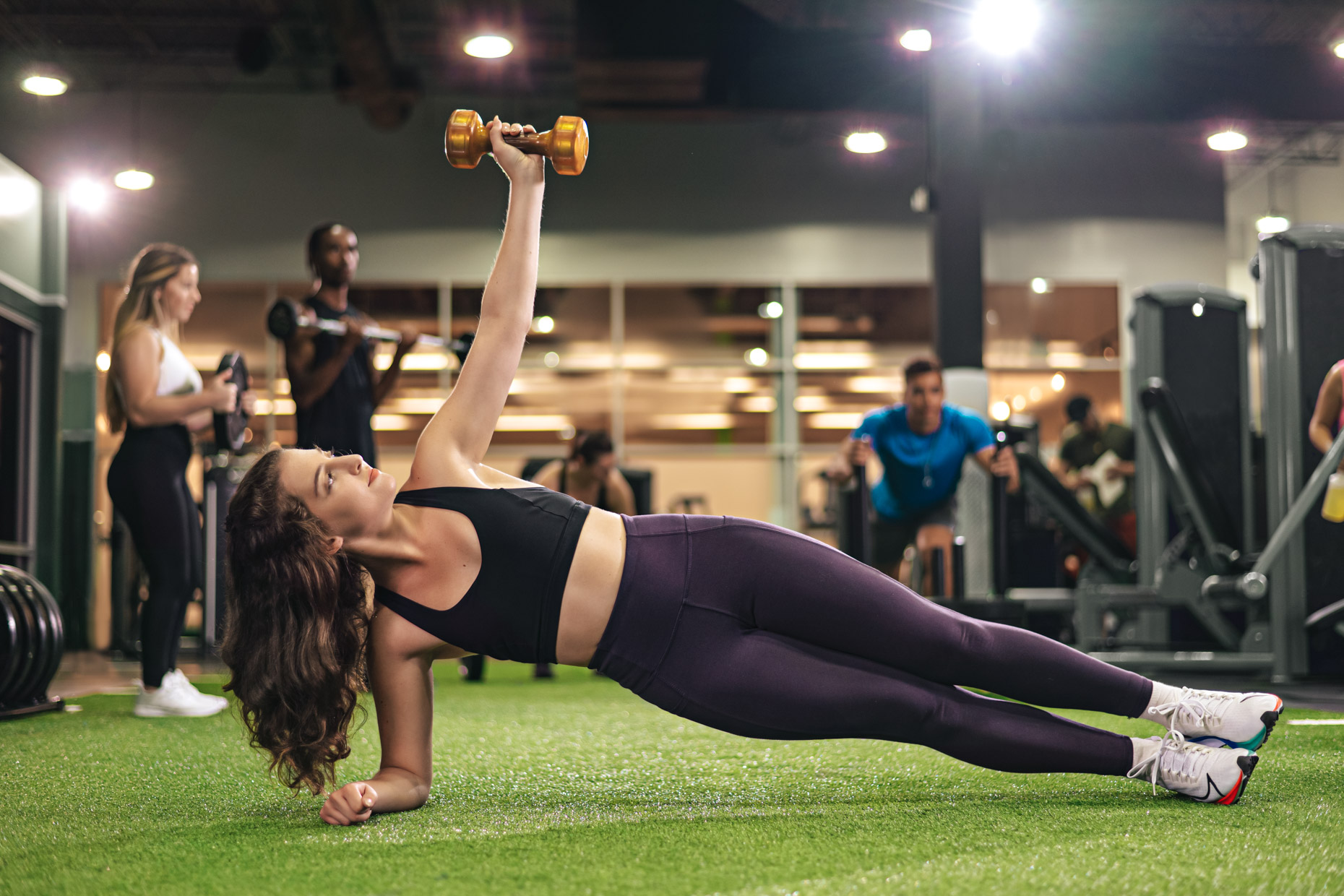 A woman in athletic wear performs a side plank while raising a dumbbell with her extended arm in a gym. Other people in the background are engaged in various fitness activities and workouts. The gym is well-lit with a spacious, modern interior.