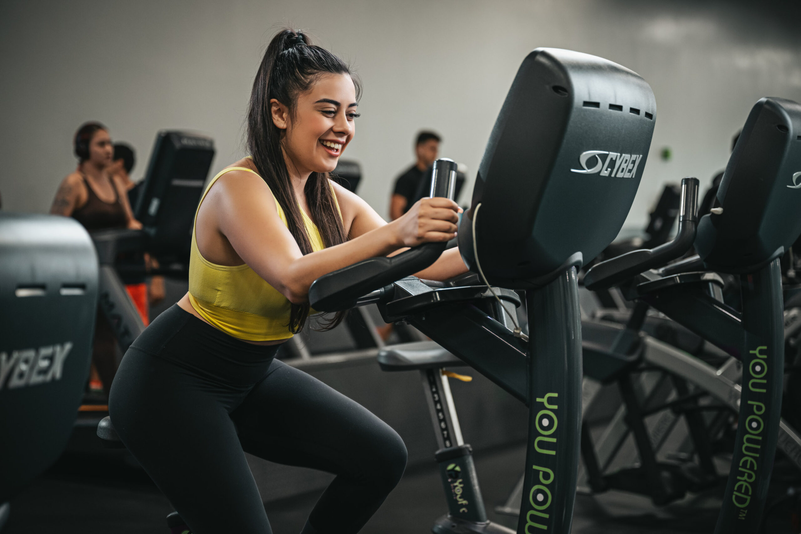 A woman in a yellow tank top and black leggings is smiling while working out on a stationary bike at a gym. Other gym-goers can be seen in the background using exercise equipment, embodying fitness for busy professionals.