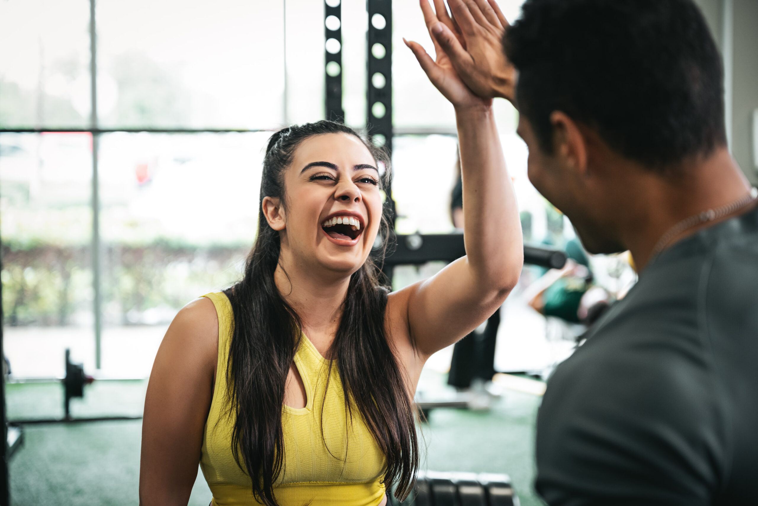 A woman in a yellow workout top and a man in a gray shirt are sharing a high-five at the gym. The woman is laughing and appears joyful. The gym setting, with its fitness equipment and window revealing greenery outside, radiates positivity—it's the perfect atmosphere for motivation hacks.