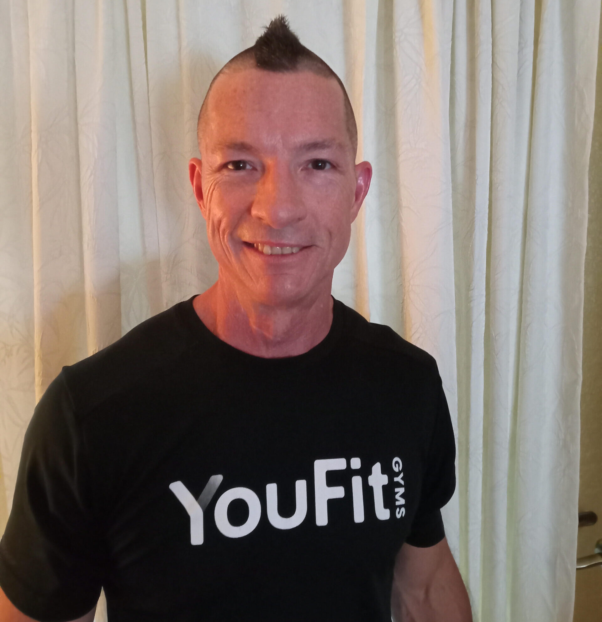 A man with a mohawk haircut smiles at the camera. He is wearing a black t-shirt with the text "YouFit GYMS" printed in white, making you think of personal training. The background consists of off-white curtains.