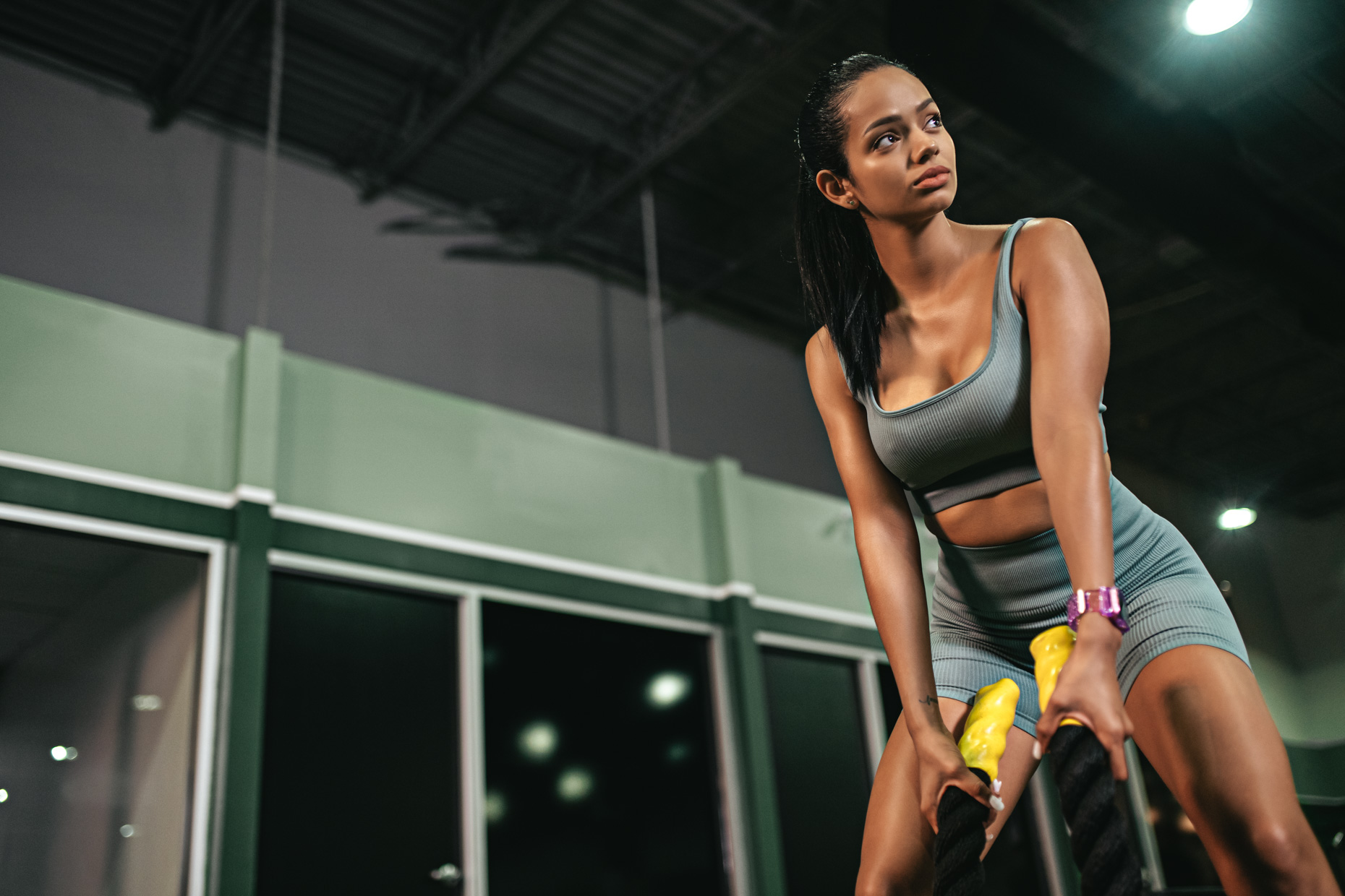 A woman dressed in a grey workout outfit exercises with battle ropes in a gym, epitomizing fall fitness. She looks focused and determined, enjoying the seasonal shift to more intense routines. The gym boasts a high ceiling and large glass windows, letting in ample natural light.