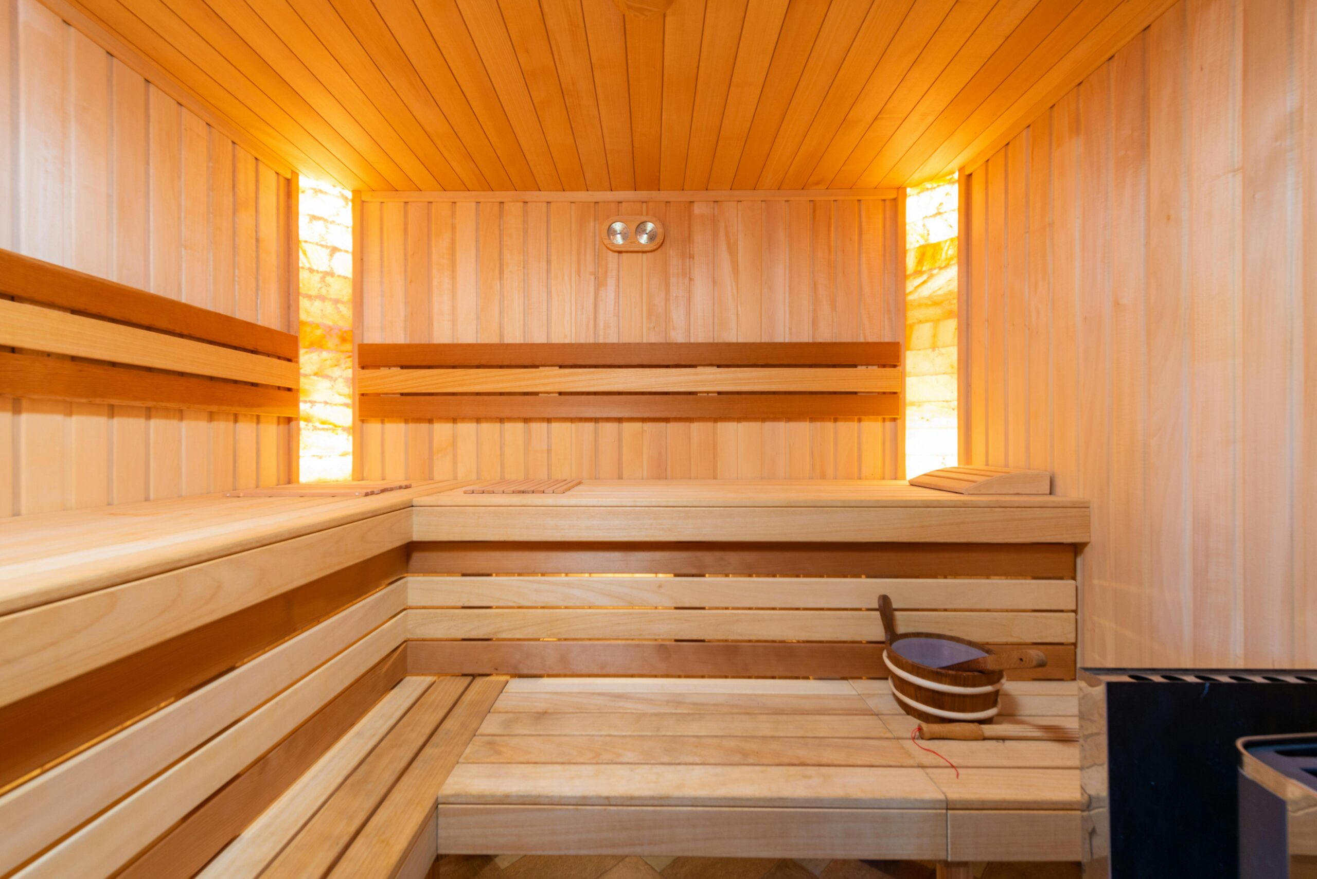 A well-lit wooden sauna interior with tiered seating along the walls. A bucket and ladle are situated on one of the lower benches. The walls and ceiling are made of light-colored wood, and a light fixture on the back wall illuminates above the seating.