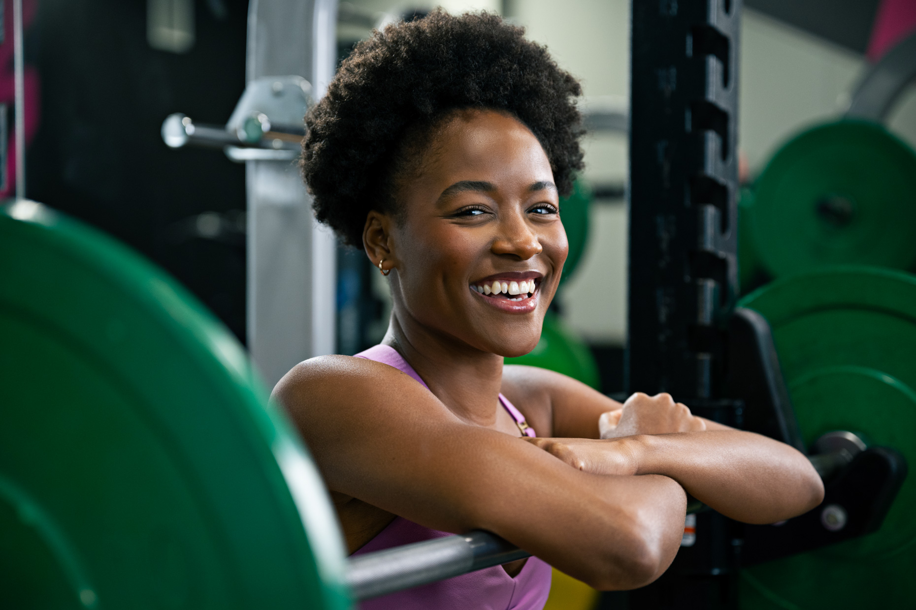 A woman, perfect for inspiring beginners, smiles as she leans on a barbell in the gym. Wearing a sleeveless pink top and rocking short, curly hair, she exudes confidence among the green weight plates and dynamic gym equipment in the energetic atmosphere.