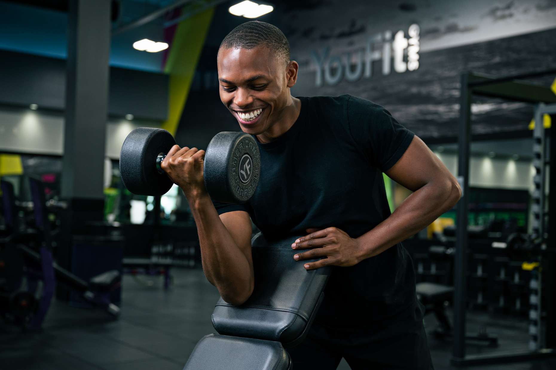 A man in a gym focuses on muscle building as he performs dumbbell curls with one arm on a workout bench. Dressed in a black t-shirt and smiling, he is surrounded by gym equipment in the background.