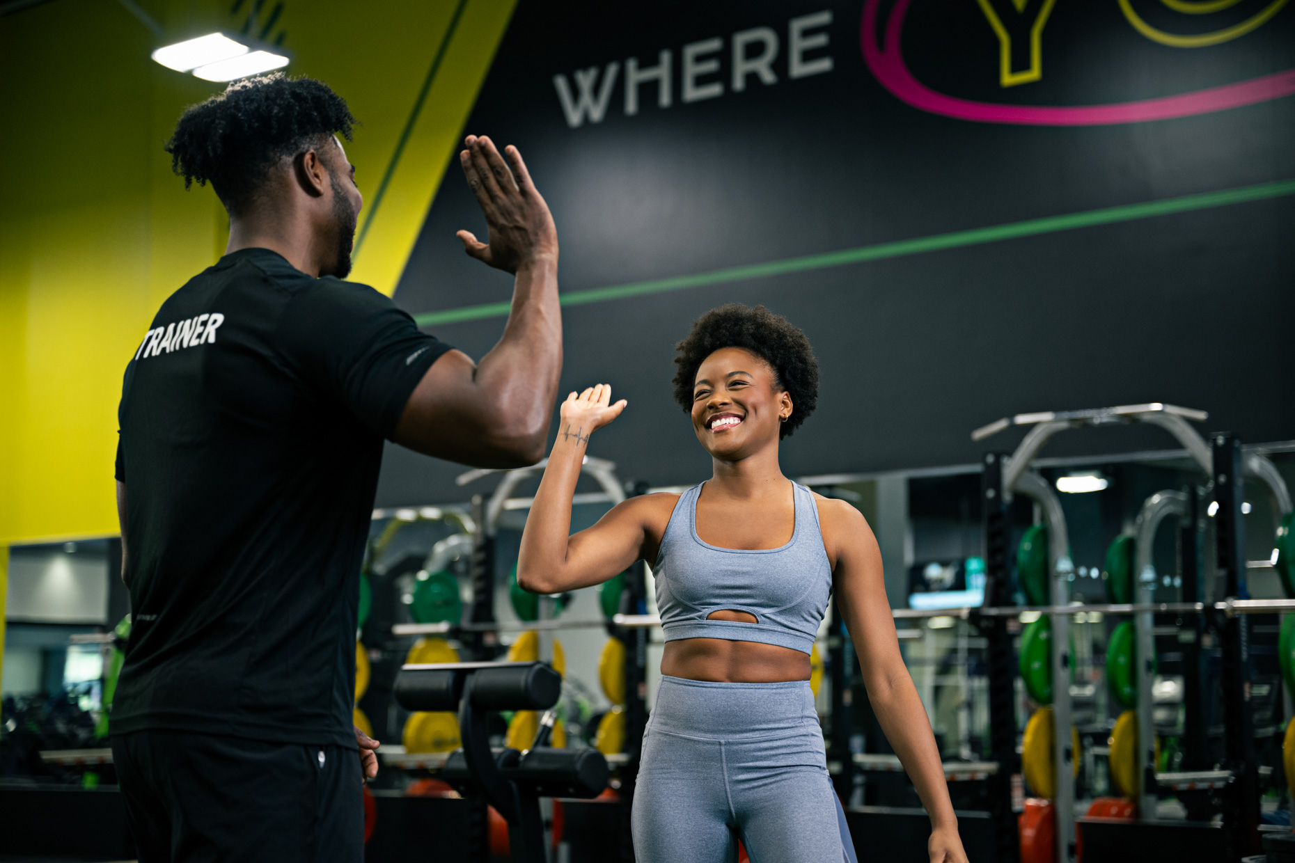 A man in a "trainer" shirt gives a high-five to a smiling woman, celebrating healthy habits in the gym. Weights and mirrors surround them, while a motivational slogan peeks from the wall in the background.