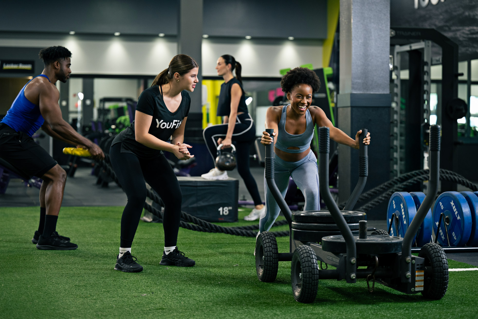 A woman is pushing a weight sled at a gym, embodying high intensity interval training as her trainer encourages her. Meanwhile, a man in the background energetically tackles the battle rope. The gym features green turf flooring and various exercise equipment.