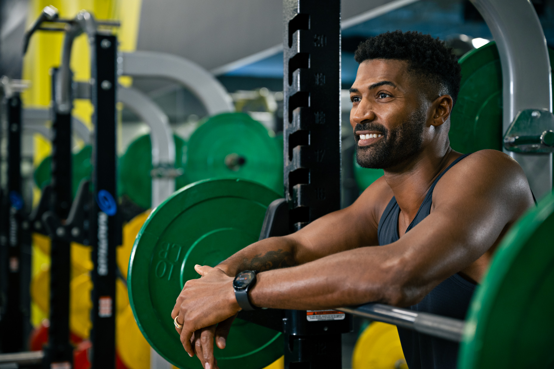 A man in a gym leans against a barbell rack, smiling confidently as if engaging in some positive self-talk. He sports a sleeveless shirt and a smartwatch, while green weights adorn the rack. Various pieces of gym equipment fill the background.