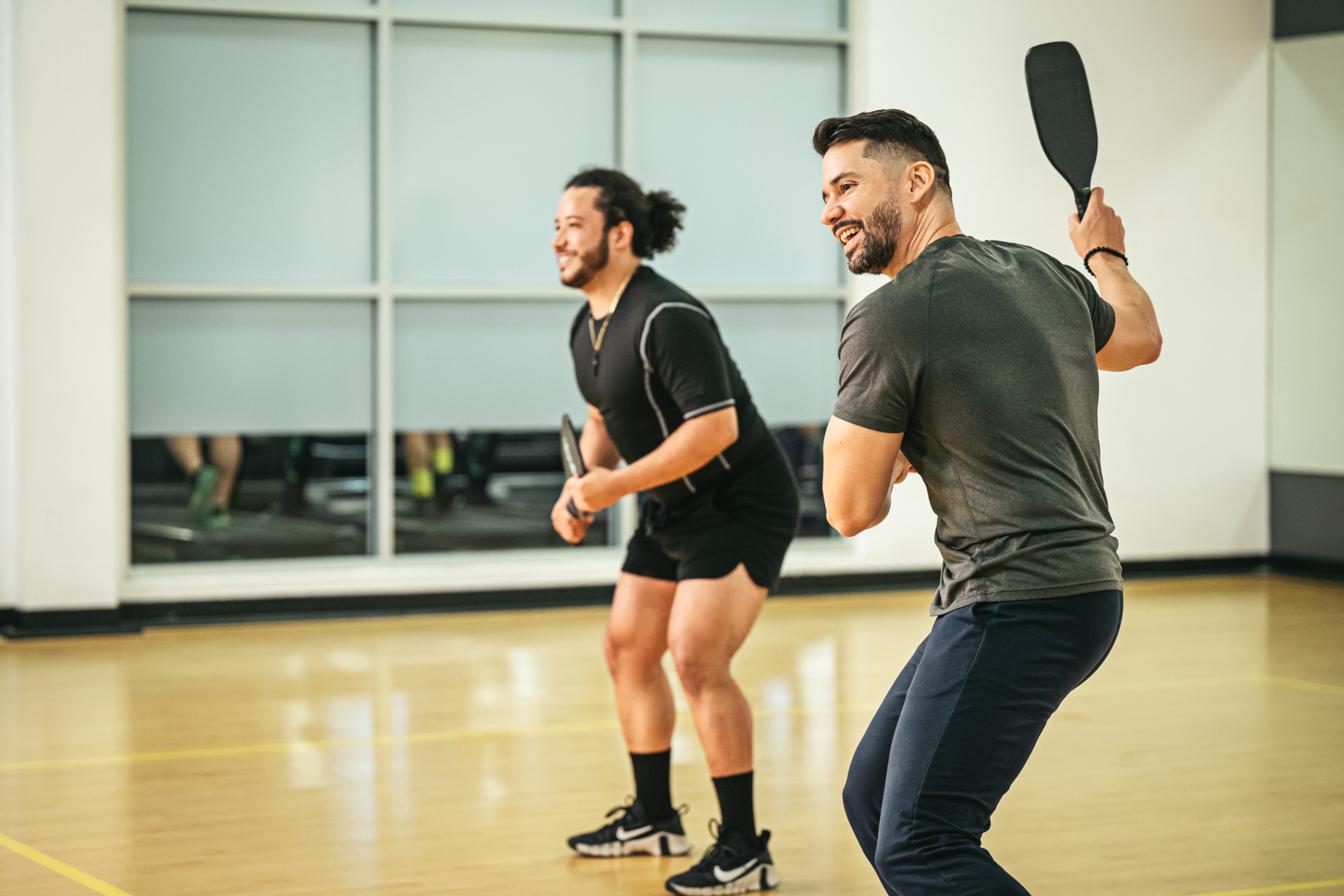 Two men playing pickleball indoors, both smiling and engaged in the game. One is preparing to hit the pickleball with his paddle while the other stands ready on the court. Large windows and gym mirrors are in the background.
