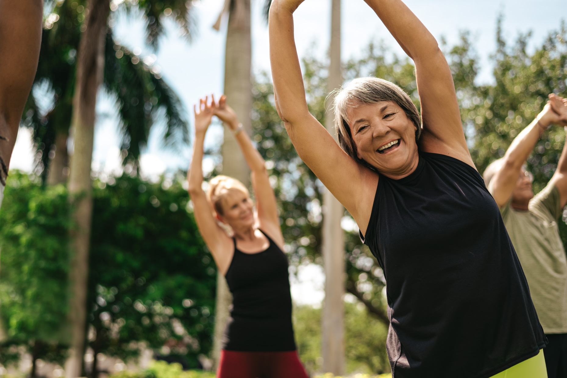 A group of adults, including an older woman smiling in the foreground, enjoy an outdoor stretch in a park with palm trees and greenery under a sunny sky.