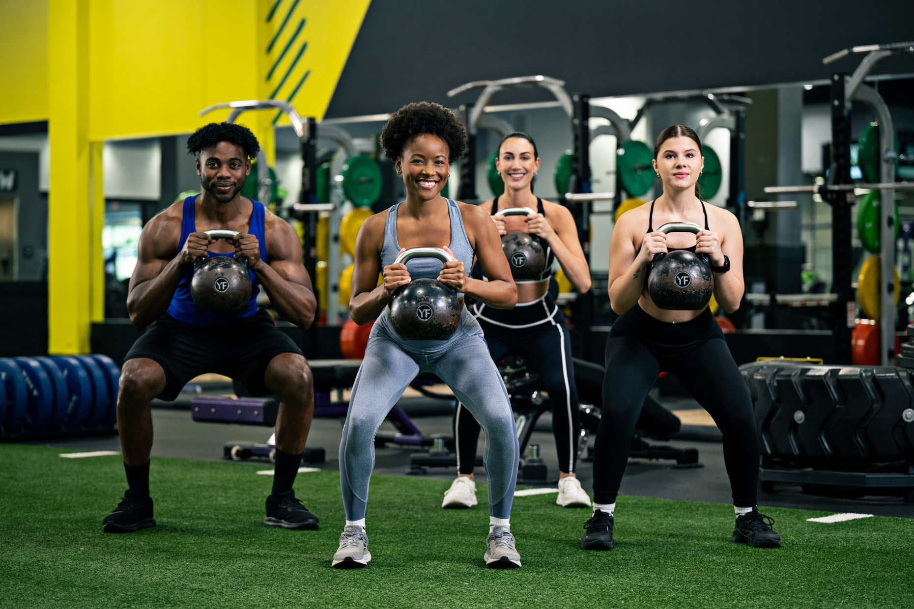 Four people exercise in a gym, smiling as they perform squats while holding kettlebells. Dressed in fitness style athletic wear, they work out together with gym equipment visible in the background.