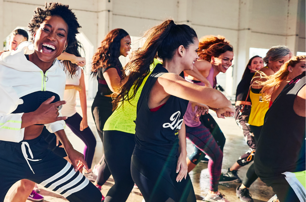 A group of people, mostly women, energetically dancing and smiling together in a bright, spacious studio. Everyone appears joyful and engaged in a lively fitness or dance class.