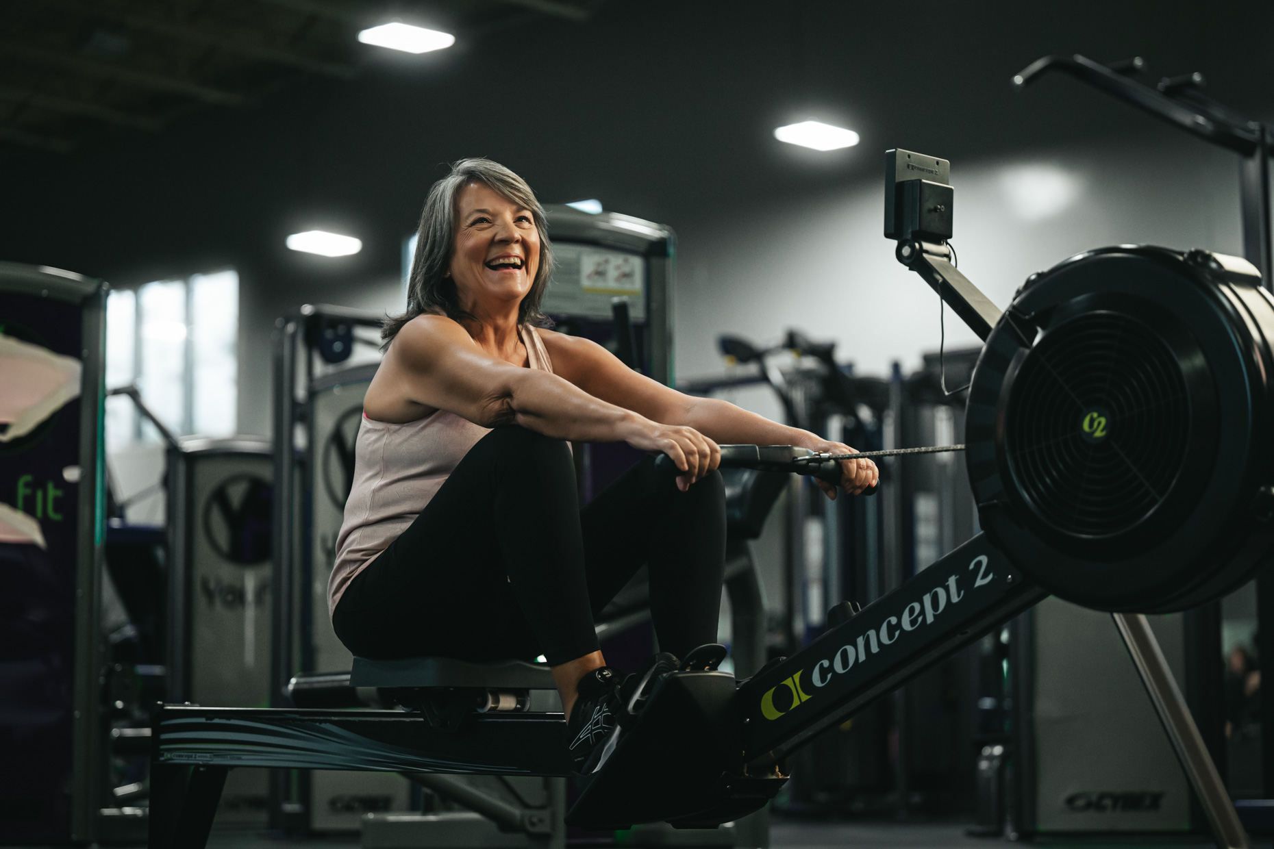 A smiling older woman engages in neuro-muscular training on a rowing machine at a well-lit gym, dressed in a light tank top and black pants, with exercise equipment visible in the background.