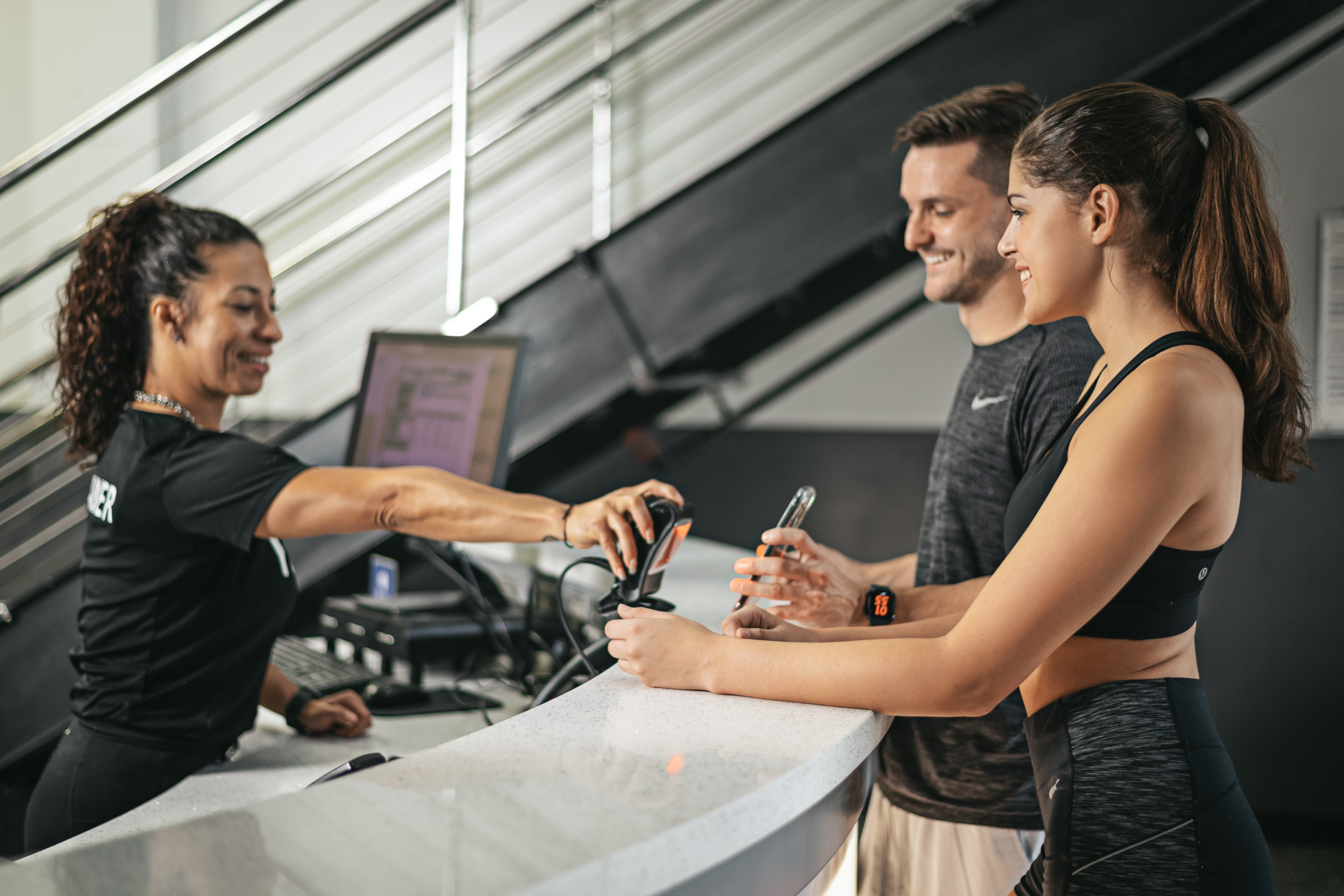 A gym receptionist scans a membership card for a smiling man and woman in athletic wear at the affordable front desk, with gym equipment and a computer in the background.
