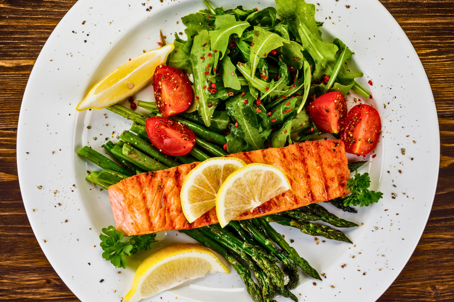 A plate with grilled salmon topped with lemon slices, served alongside asparagus, arugula salad with cherry tomatoes, and extra lemon wedges—an ideal post-workout meal garnished with herbs and black pepper.