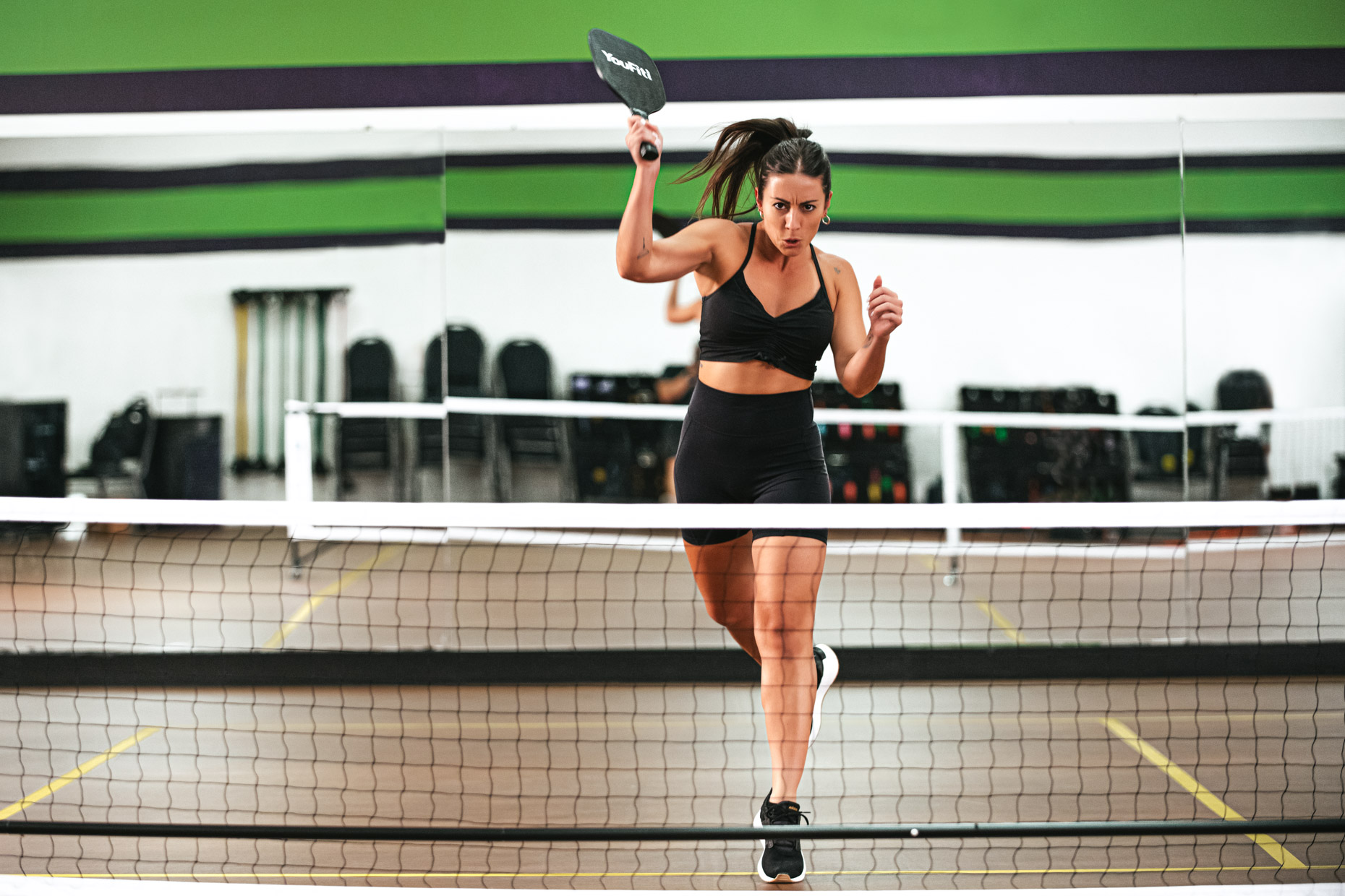 A woman in athletic wear plays pickleball indoors, preparing to hit the ball with her paddle while stepping forward near the net on a pickleball court.