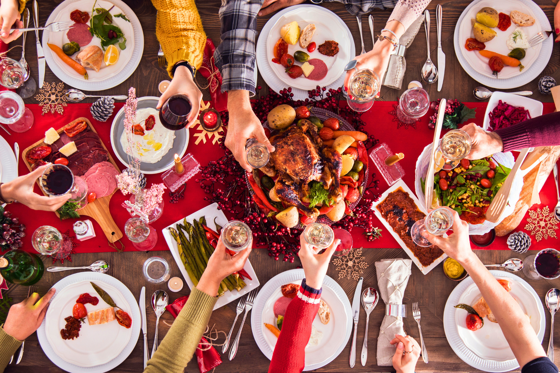 A festive table set for a holiday meal, with people raising glasses in a toast around a roasted turkey, vegetables, wine, and mindful eating for the holidays, all viewed from above.