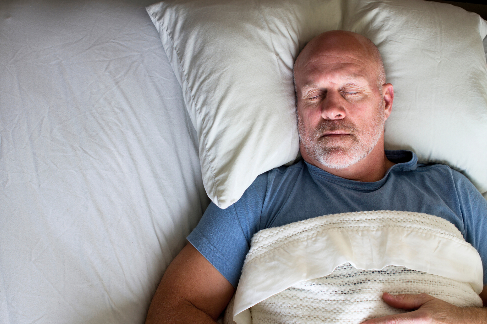 A bald, middle-aged man with a gray beard is lying on his back in bed, eyes closed, following a healthy sleep schedule as he rests peacefully on a white pillow, wearing a blue t-shirt and covered with a white blanket.