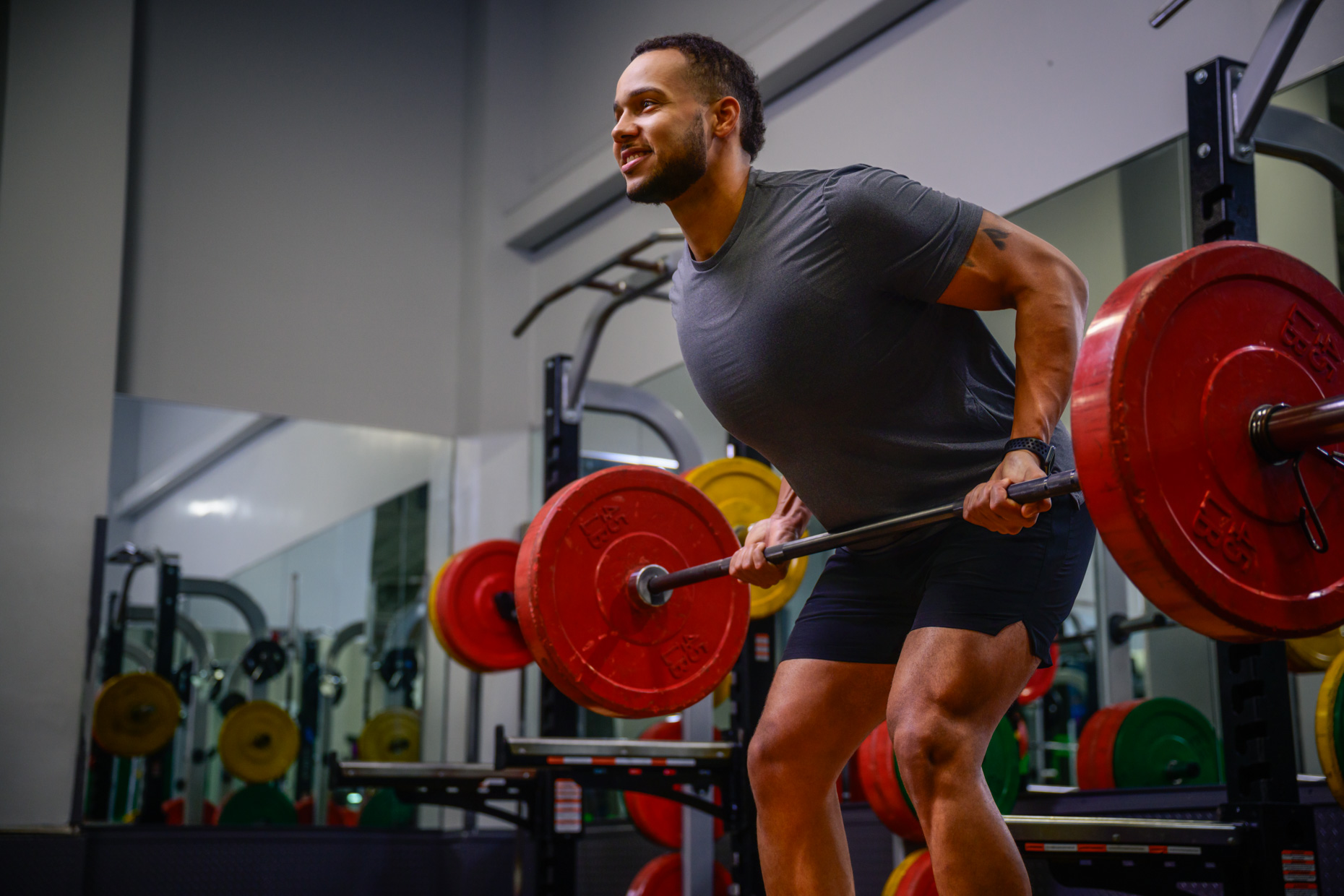 A man in athletic wear lifts a barbell with red weight plates in a gym, performing a bent-over row to promote hypertrophy. He is smiling and surrounded by various weightlifting equipment and mirrors.