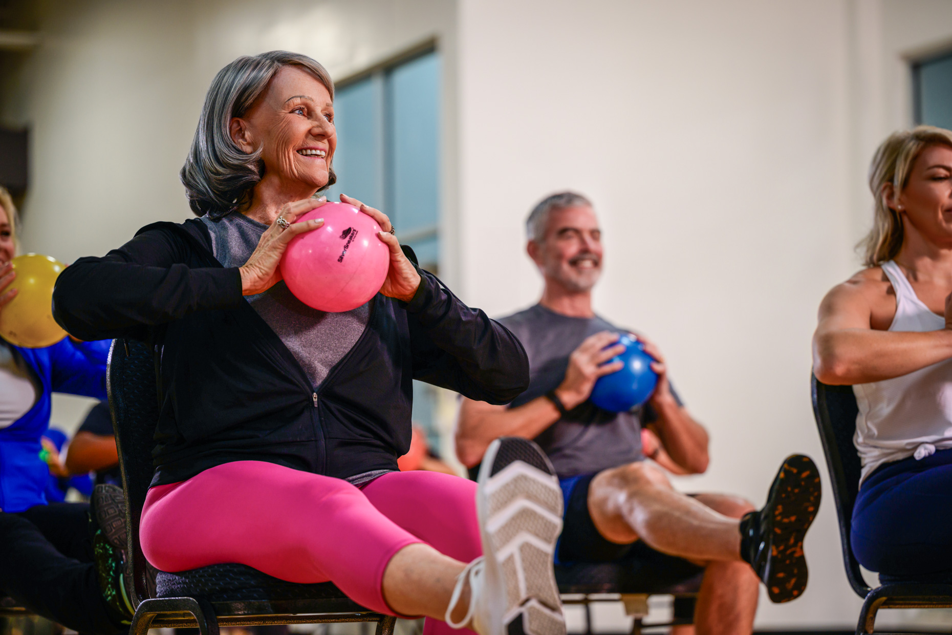 A group of older adults exercise in a gym after 60, sitting on chairs and lifting colorful balls while smiling; a gray-haired woman in pink leggings is in the foreground.
