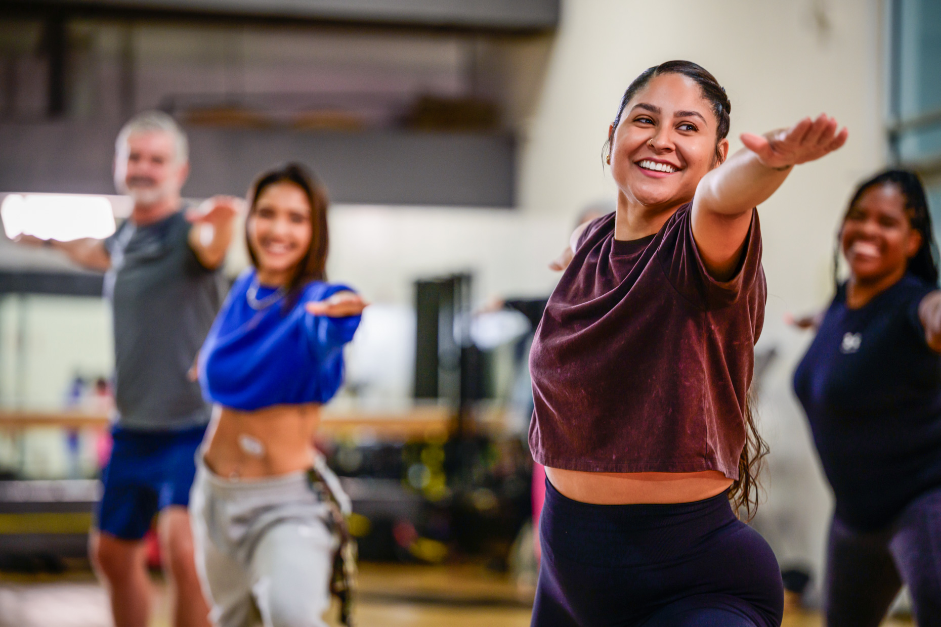 A group of four adults exercising together in a fitness studio, smiling and holding their arms outstretched as they perform a workout that blends lifting and yoga, with a focus on a cheerful woman in the front.
