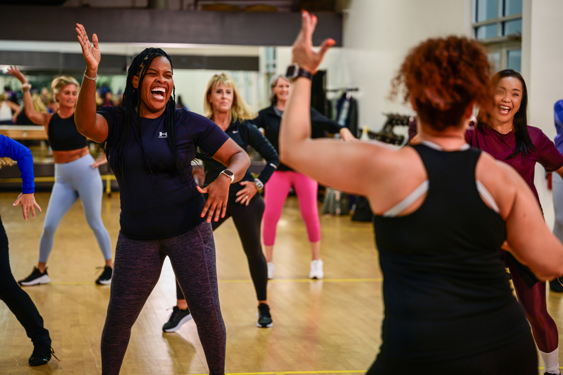 A group of women smile and dance energetically in a fitness studio, following an enthusiastic instructor, as they shake off holiday stress. The room features wooden floors, mirrors, and exercise equipment in the background.