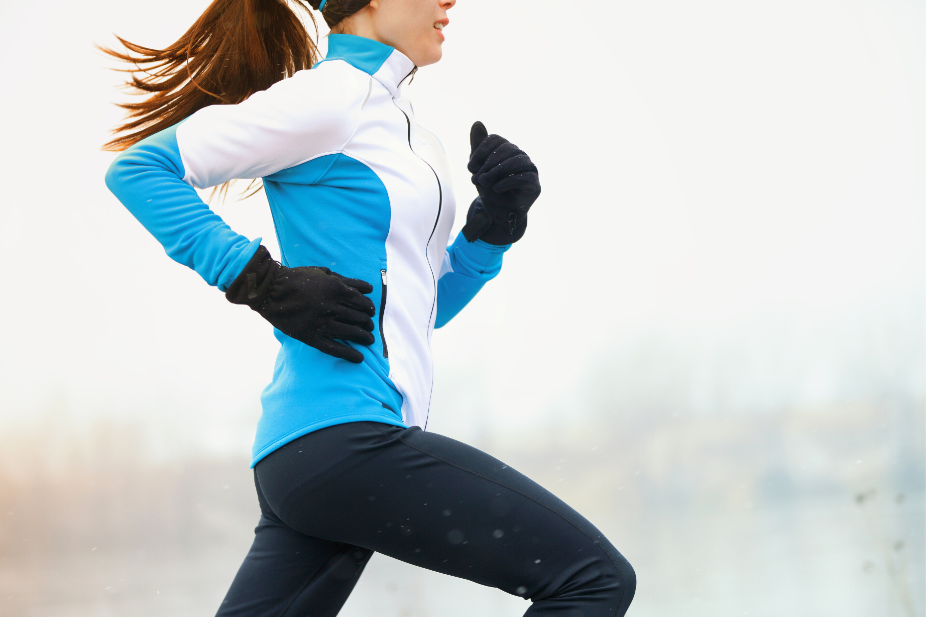 A woman in a blue and white jacket and black gloves is running outdoors on a cold day, embracing healthy cold weather exercise. The background is blurred, suggesting motion, and her hair is tied back.