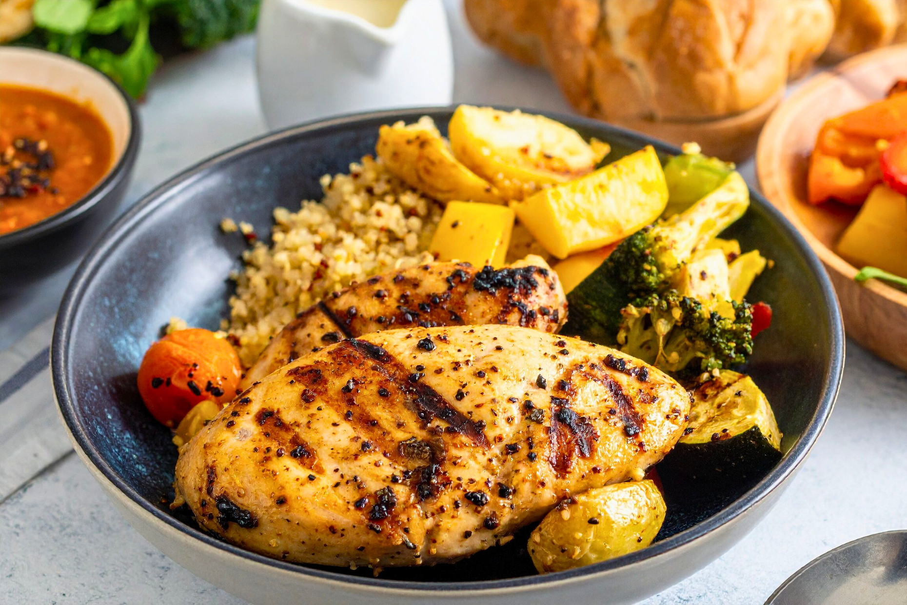 A meal prep bowl with grilled chicken breast, quinoa, and roasted vegetables like yellow squash, broccoli, and tomatoes, served on a table with bread and sauce in the background.