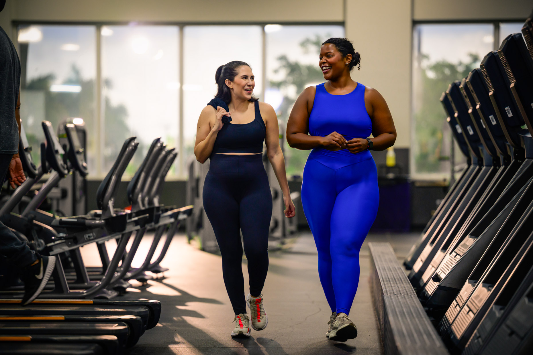 Two women in athletic wear walk and chat together in a gym, smiling as they pass by treadmills and exercise equipment, getting back on track with their fitness goals in the bright, natural light from large windows.
