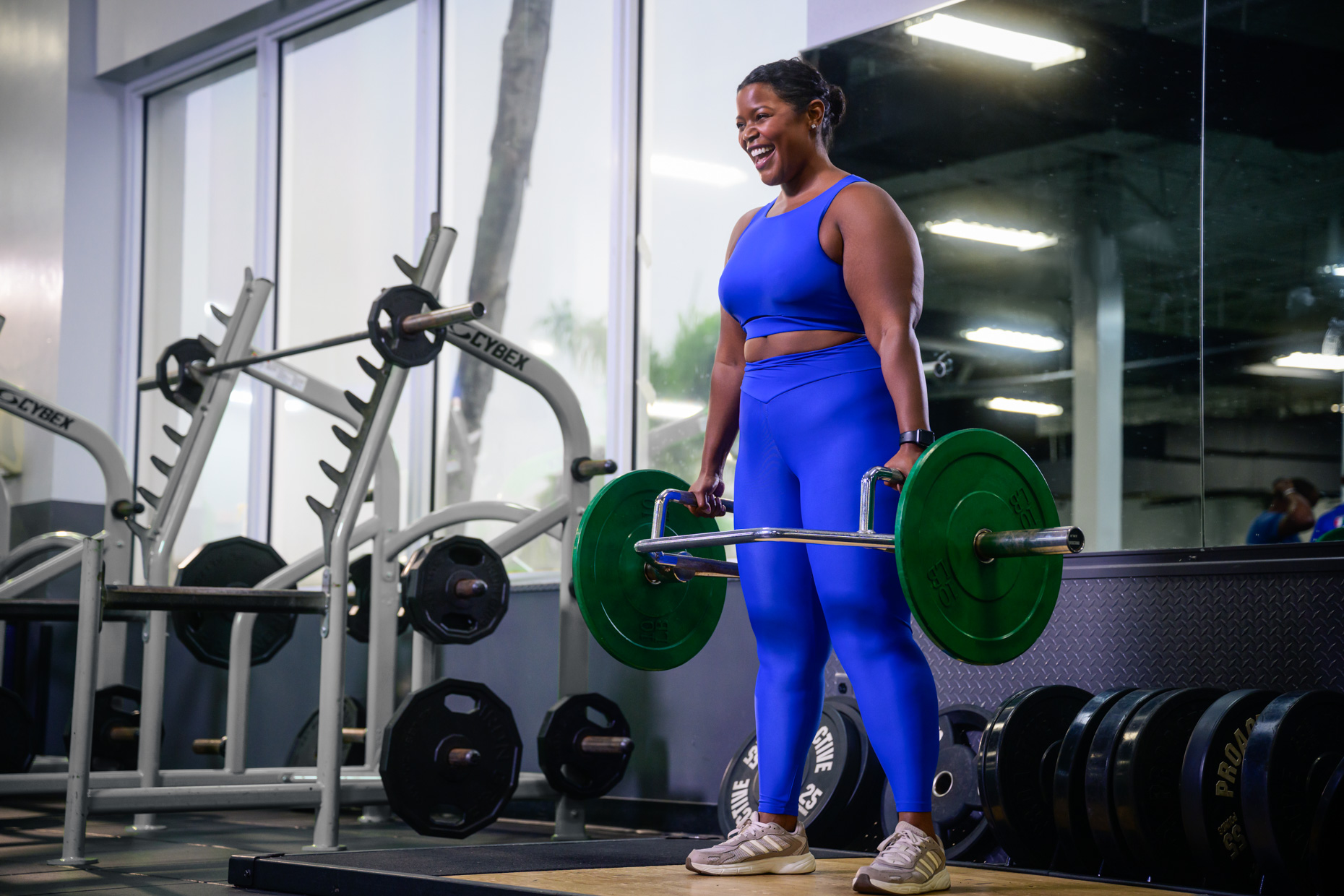 A woman in a bright blue athletic outfit smiles while lifting a loaded trap bar in a 2026 gym, surrounded by weight plates and gym equipment.